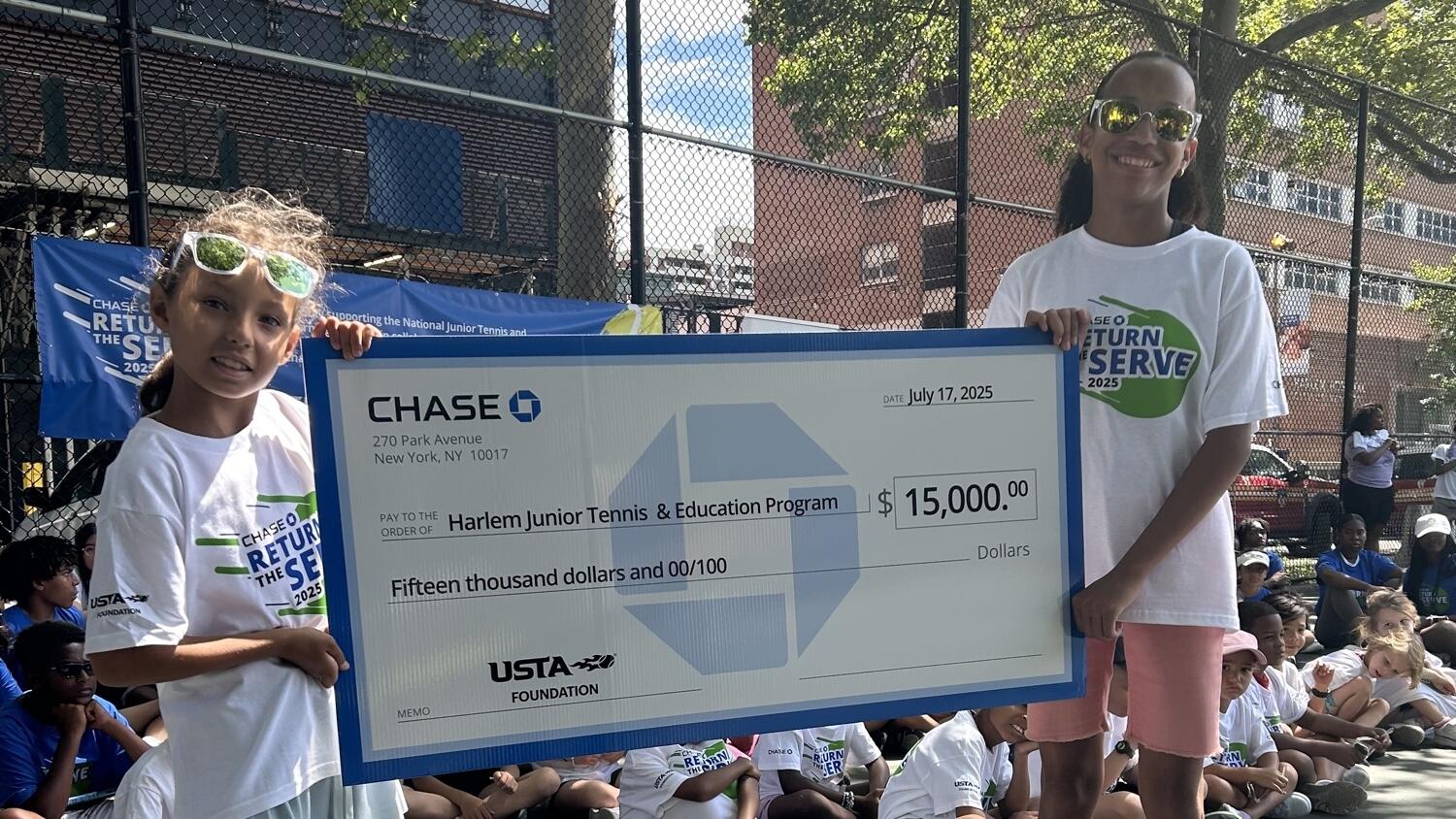Two young tennis players hold a large ceremonial check for $15,000 made out to the Harlem Junior Tennis & Education Program from the USTA Foundation at an outdoor tennis court event.