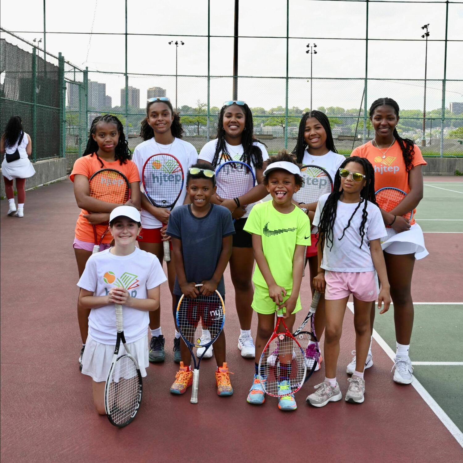 A group of young tennis players pose together on an outdoor court at Riverbank State Park, holding tennis rackets and wearing HJTEP shirts. The older student-athletes stand in the back row while younger children kneel in front, all smiling at the camera with the Manhattan skyline visible through the court's chain-link fence in the background.RetryClaude can make mistakes. Please double-check responses.