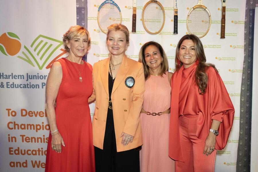 Four women, including Chris Evert, in coordinating coral and orange formal attire pose together at a fundraising event in front of a step-and-repeat banner for Harlem Junior Tennis & Education Program. The backdrop features the organization's logo with tennis racket graphics and text about developing champions in tennis, education and wellness.