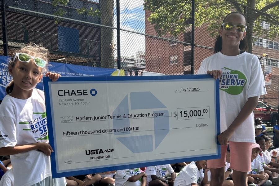 Two young tennis players hold a large ceremonial check for $15,000 made out to the Harlem Junior Tennis & Education Program from the USTA Foundation at an outdoor tennis court event.