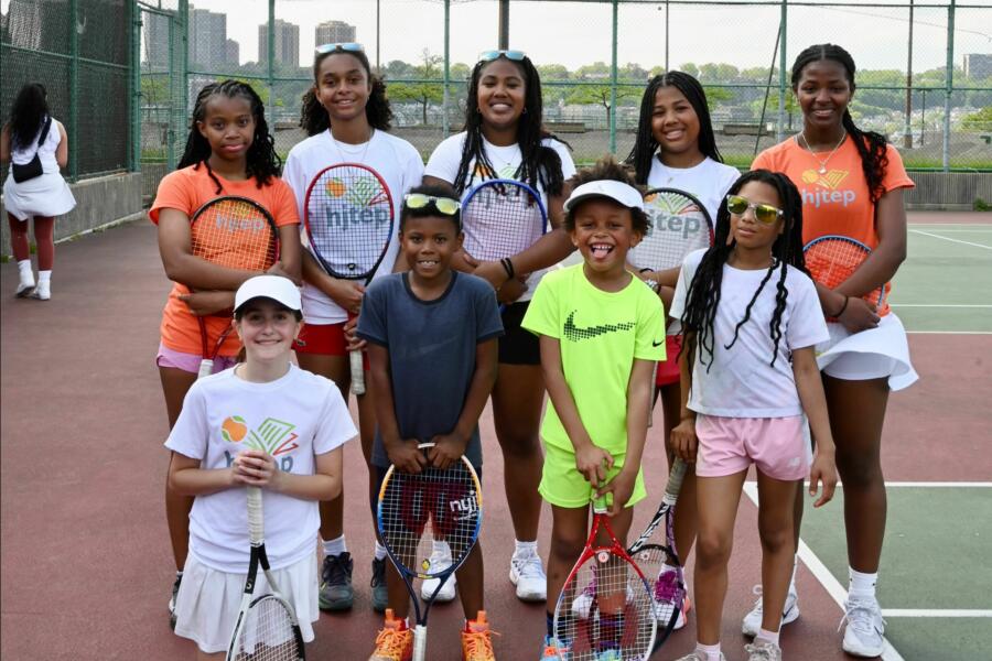 A group of young tennis players pose together on an outdoor court at Riverbank State Park, holding tennis rackets and wearing HJTEP shirts. The older student-athletes stand in the back row while younger children kneel in front, all smiling at the camera with the Manhattan skyline visible through the court's chain-link fence in the background.RetryClaude can make mistakes. Please double-check responses.